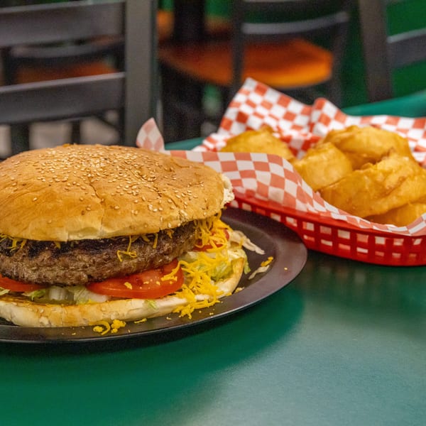 Large burger with cheese, lettuce, and tomato, served with a side of onion rings in a red basket.