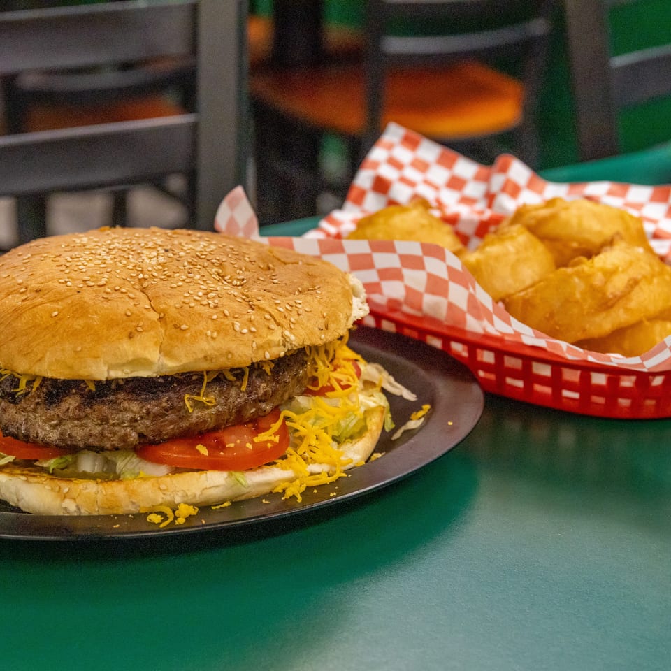 Large burger with cheese, lettuce, and tomato, served with a side of onion rings in a red basket.