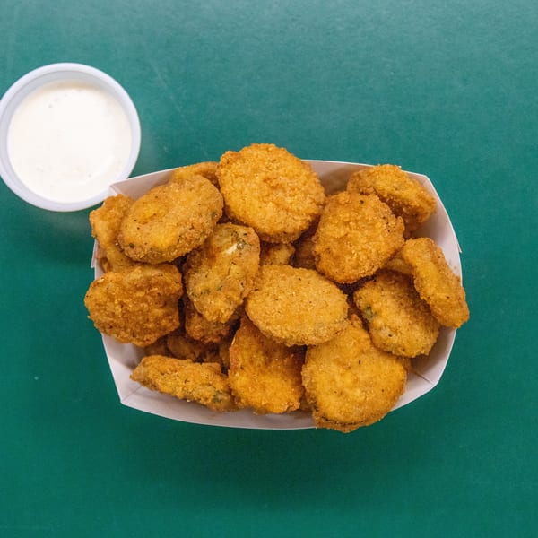 Fried pickles in a paper container with a side of white dipping sauce on a green surface.