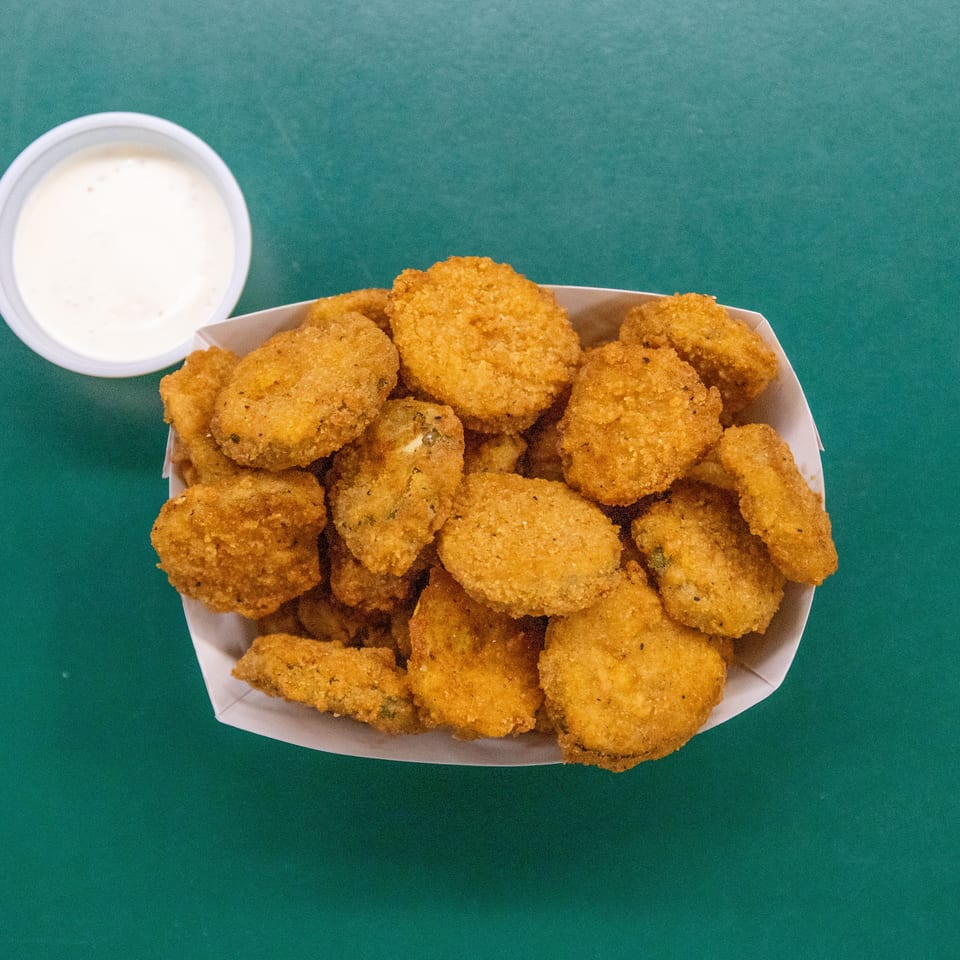 Fried pickles in a paper container with a side of white dipping sauce on a green surface.
