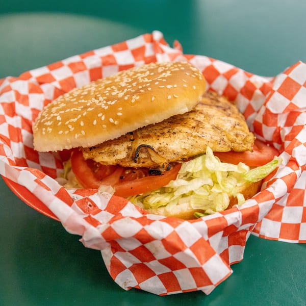 Grilled chicken sandwich with lettuce, tomato, and sesame bun in a red checkered paper liner.
